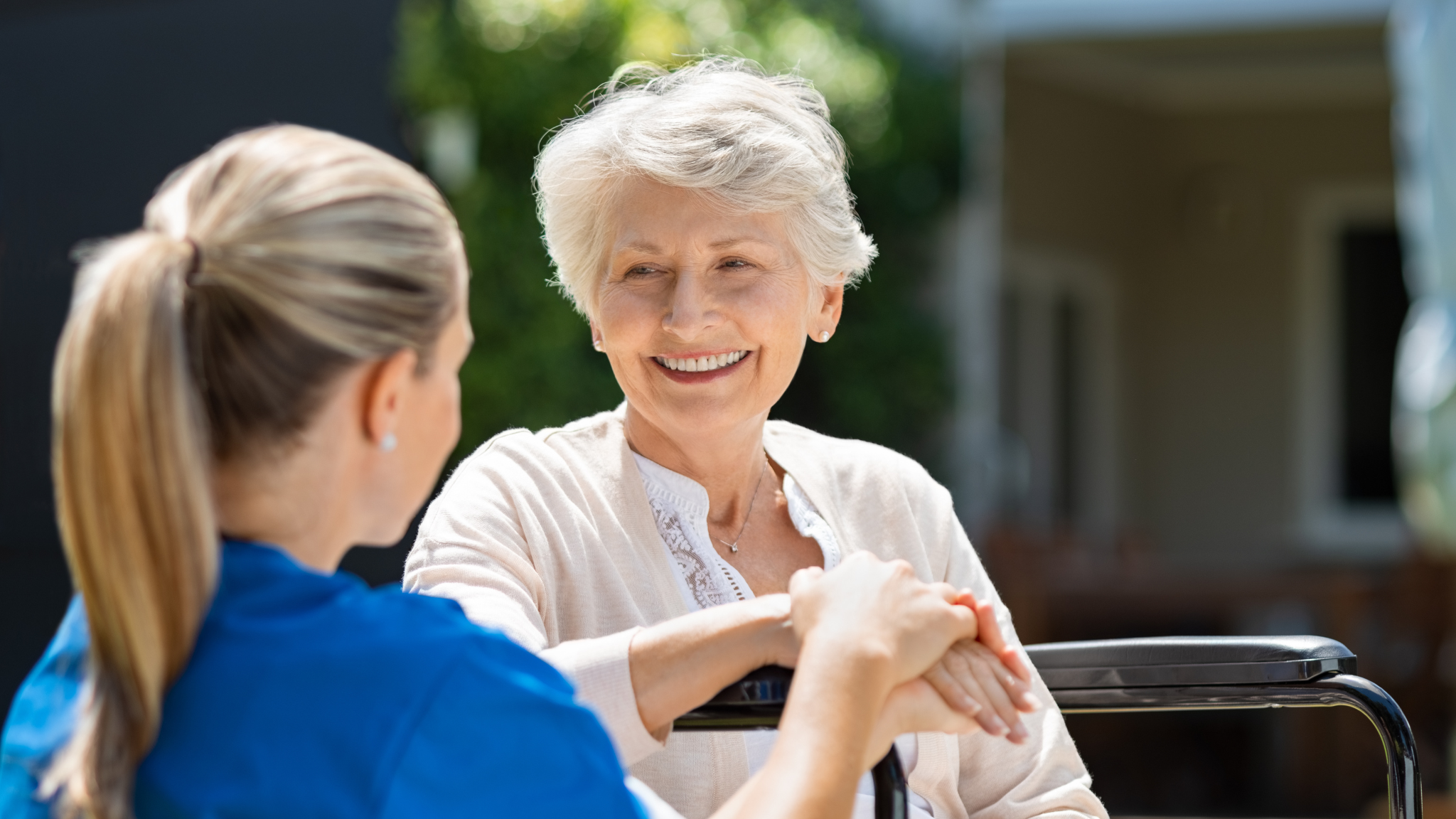In Maple Ridge BC, a smiling, older woman sits in a wheelchair, her hands gently held by a healthcare worker in a blue uniform.