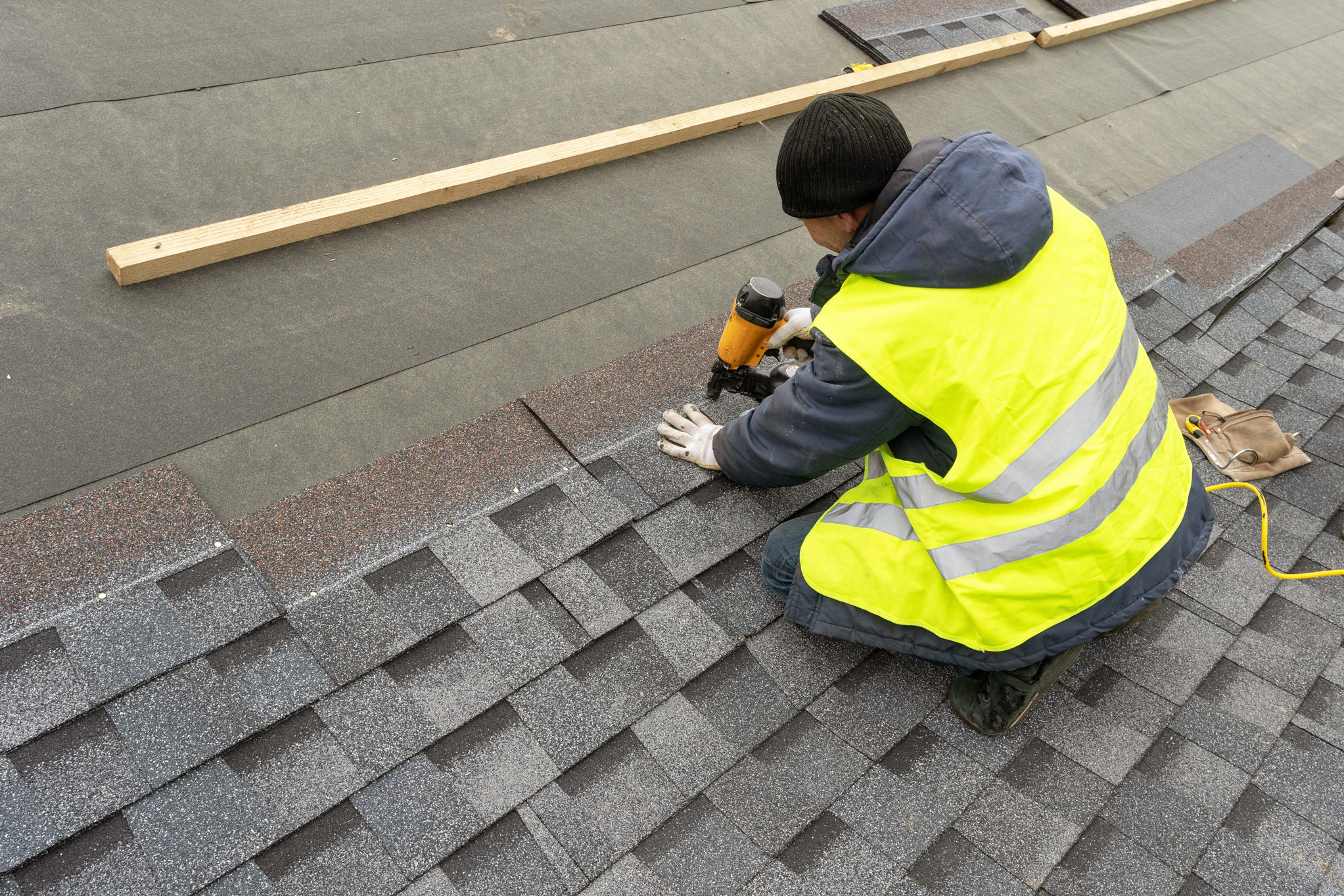 In Maple Ridge BC, a worker wearing a neon safety vest uses a nail gun while installing roofing shingles.