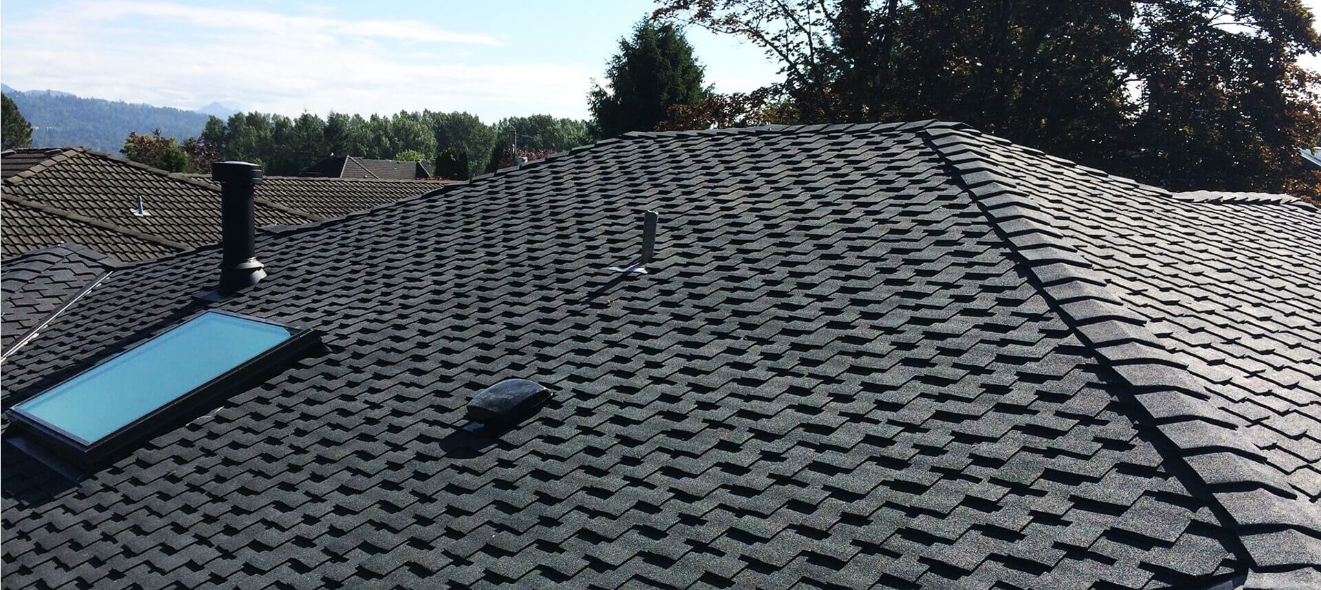 A Maple Ridge BC roof with dark grey shingles, a skylight, and vents. Other rooftops and trees appear in the background under a blue sky.