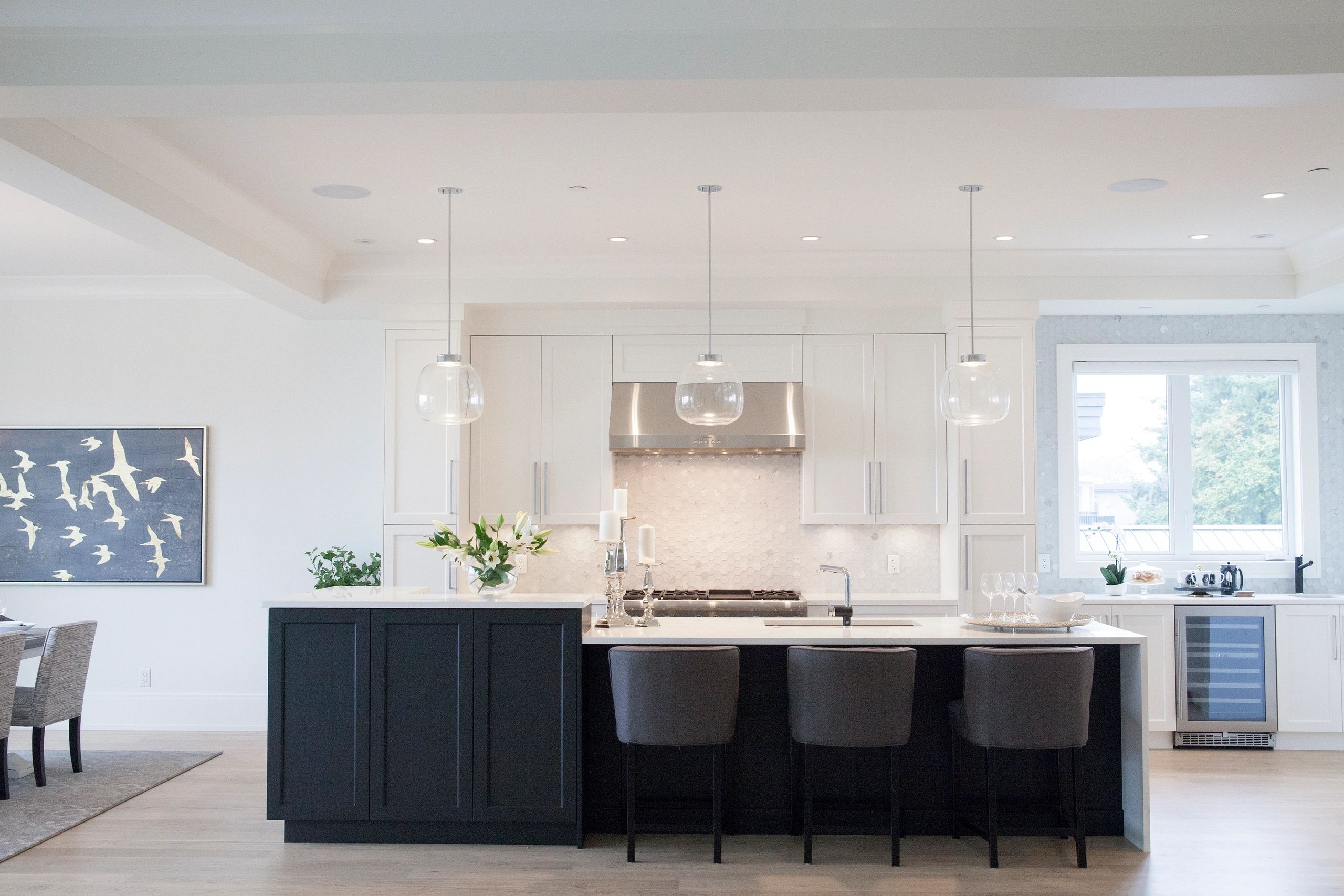 A well-lit kitchen in Maple Ridge BC features a central island with bar stools, pendant lights, and a dining area visible in the background.