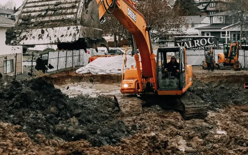 A Deere excavator digs into the earth, with another worker nearby, near a Valroc sign in Maple Ridge BC.