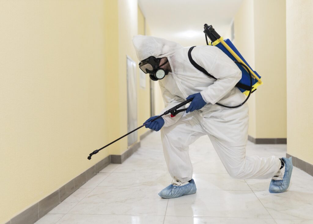 In Maple Ridge BC, a person in full protective gear, including a mask, gloves, and suit, sprays the base of a hallway wall using a tank and wand apparatus.