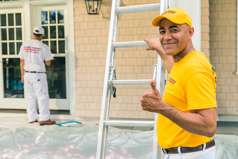 A painter in Maple Ridge BC, wearing a company shirt and hat, gives a thumbs up next to a ladder, while another painter works on trim.