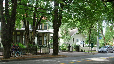 Street scene in Maple Ridge BC, featuring a couple with a bicycle near a vintage building shaded by trees. A house with a gated fence and a parked car are visible further down the street.